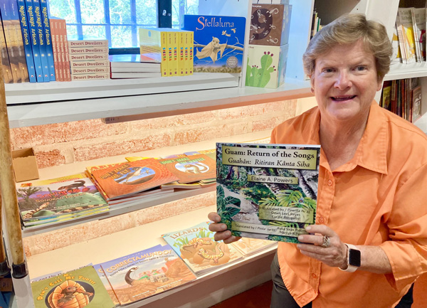 Elaine, holding her newest book in front of a glass case with her other picture books on display.