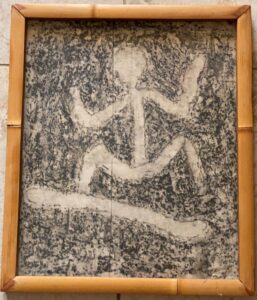 A framed black and white petroglyph rubbing that depicts a human figure surfing. 