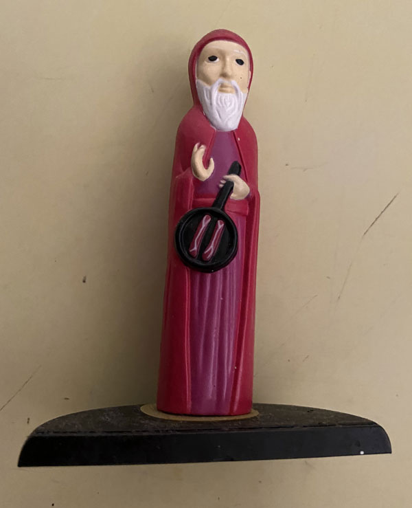 A small statuette of St. Anthony, he wears a long hooded red robe and holds a skillet with two slices of bacon.