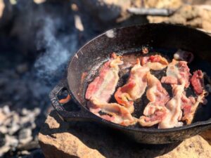 Four strips of bacon cooking in a cast iron skillet over an outdoor camp fire.