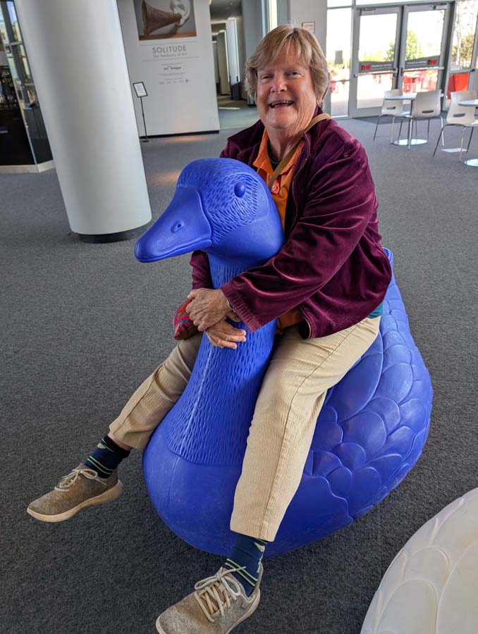 Elaine on a large, blue plastic goose. Her feet are dangling over its sides.